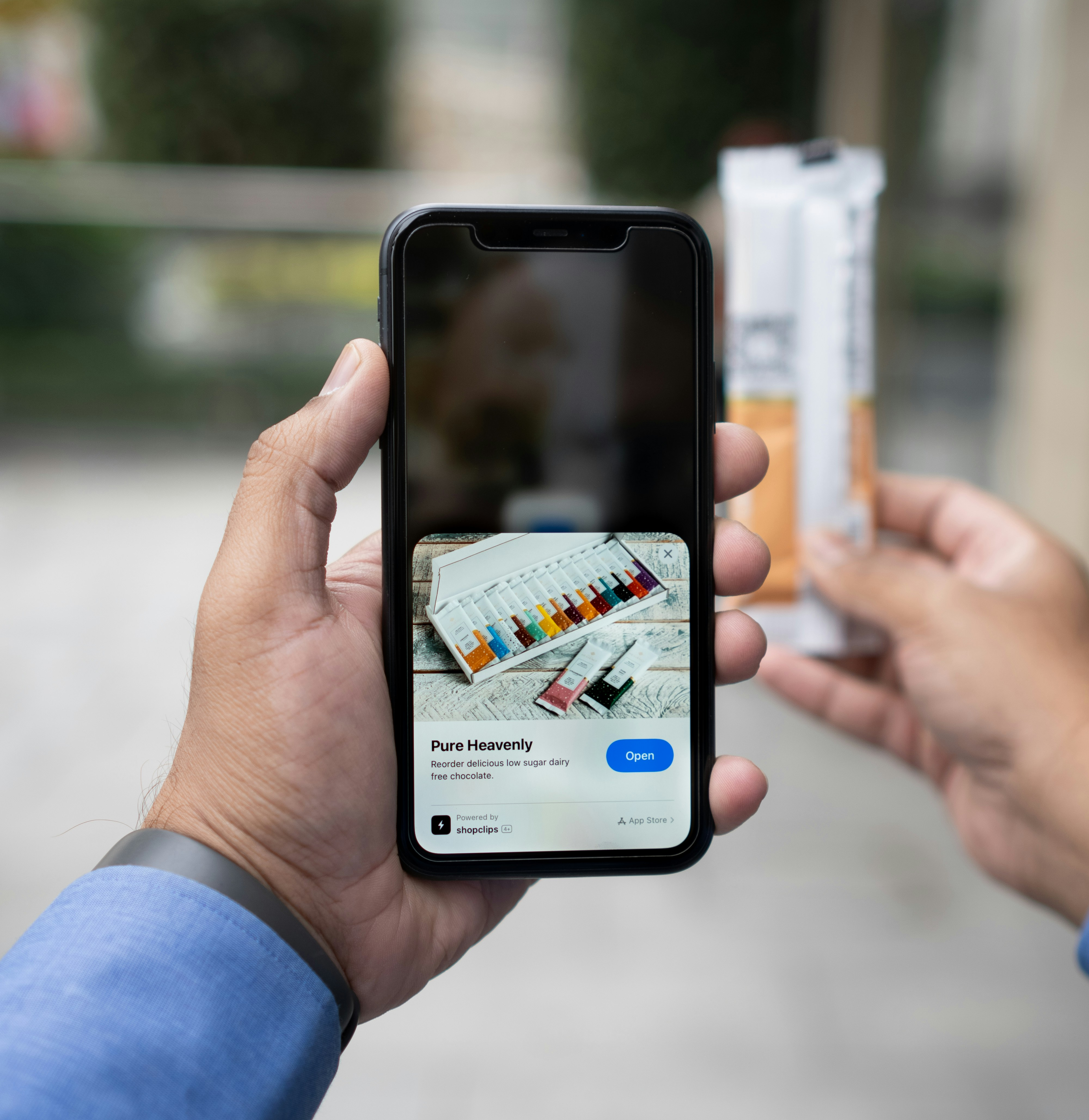 A hand holds a smartphone showing an online shopping app for chocolate, while scanning a product held in the other hand outdoors.