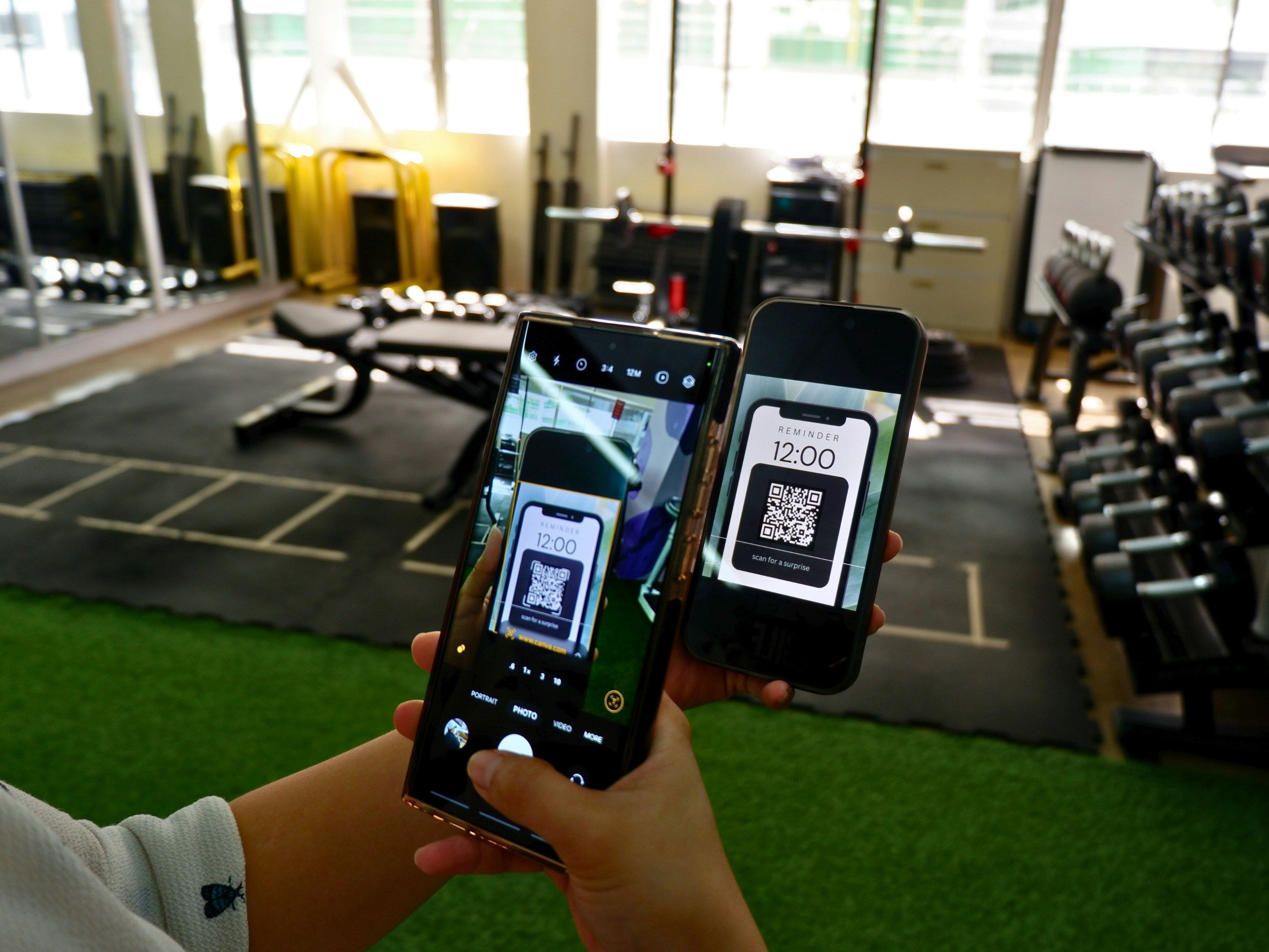 A person holds a smartphone displaying a 12:00 reminder with a QR code, while taking a photo of it using another phone inside a gym with workout equipment in the background.